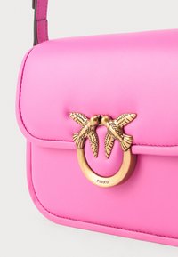 Borsa a tracolla in pelle rosa con design strutturato, caratterizzata da un'accogliente accentuazione dorata di un uccello e hardware circolare con il nome del marchio "PINKO" inciso.