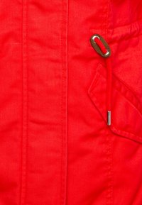 Veste en tissu rouge avec une texture lisse, dotée d'une poche latérale et d'un œillet en métal contrastant avec un cordon rouge pour le réglage.