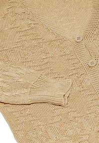 Beige stickad kappa med ett strukturerat mönster, V-hals och stora knappar. Tyget har ett andningsbart och lätt utseende.