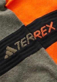Tessuto grigio e arancione con righe verticali, accentuato da dettagli neri. Il marchio "TERREX" è ricamato in arancione e marrone sopra un simbolo del logo.