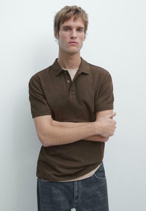 Polo marron à manches courtes avec col, deux boutons et une finition texturée, associé à un jean clair. Le mannequin pose avec les bras croisés.