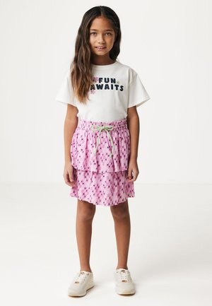 T-shirt bianco con la scritta "DIVERTIMENTO IN ARRIVO" in blu e design floreale. Gonna rosa a strati con motivo geometrico e cordoncino verde. Sneaker bianche.