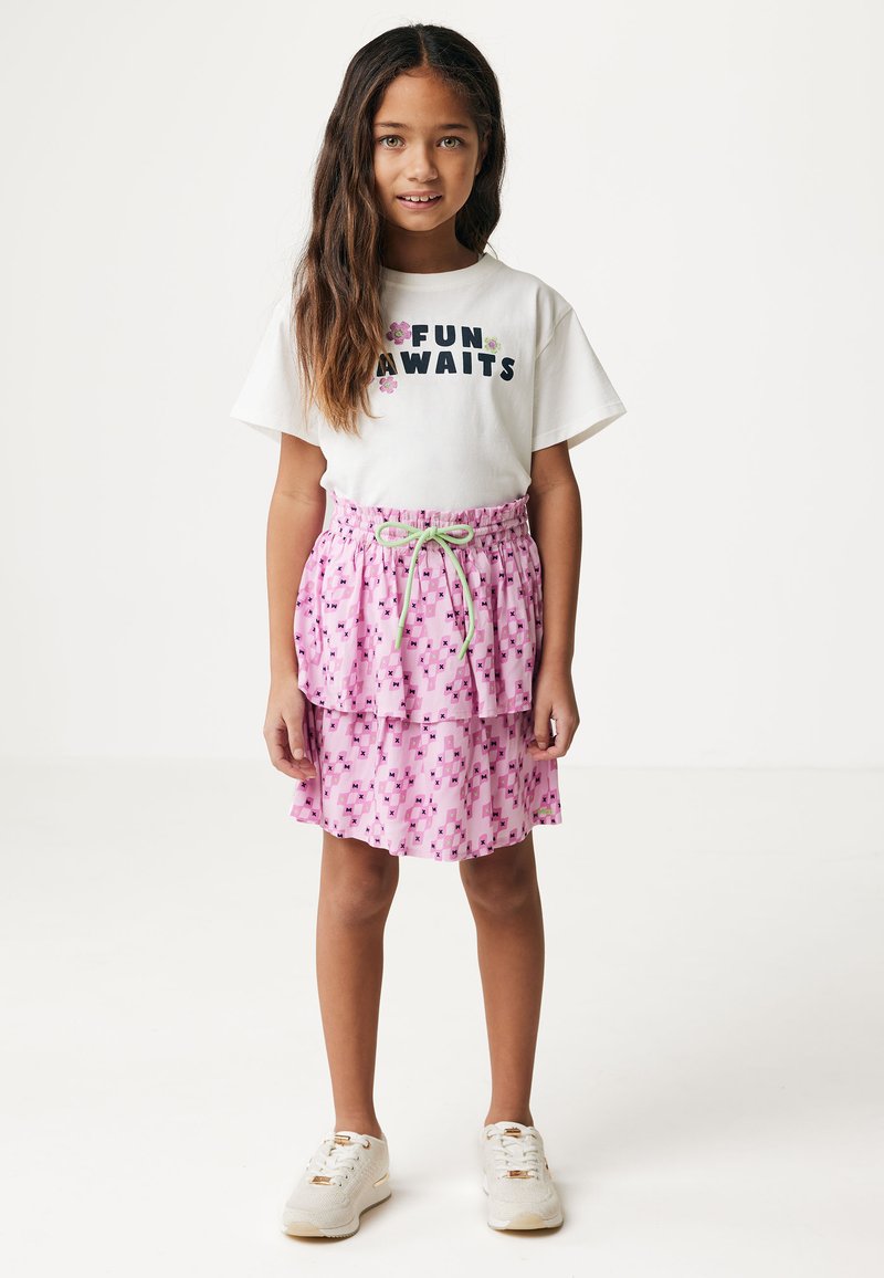 T-shirt bianco con la scritta "DIVERTIMENTO IN ARRIVO" in blu e design floreale. Gonna rosa a strati con motivo geometrico e cordoncino verde. Sneaker bianche.