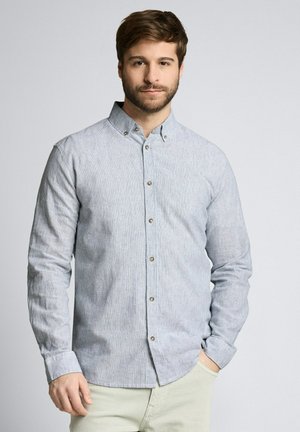 Mann mit braunen Haaren und Bart, der ein hellblau gestreiftes Hemd mit Knöpfen und beige Hosen trägt, steht mit einer Hand in der Tasche.