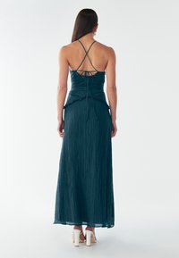 Abito maxi teal con corpetto aderente e orlo arricciato. Presenta spalline incrociate sul retro e tessuto testurizzato. Indossato con tacchi bianchi.