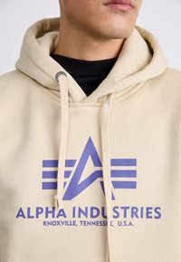 Bežinė džemperis su purpuriniu logotipu, kuriame yra geometrinis dizainas. Turi raištinį gaubtą ir matomus siuvimo detalės.