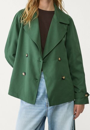 Femme portant une veste verte croisée à grands revers et boutons en écaille, associée à un jean bleu clair et un haut foncé.