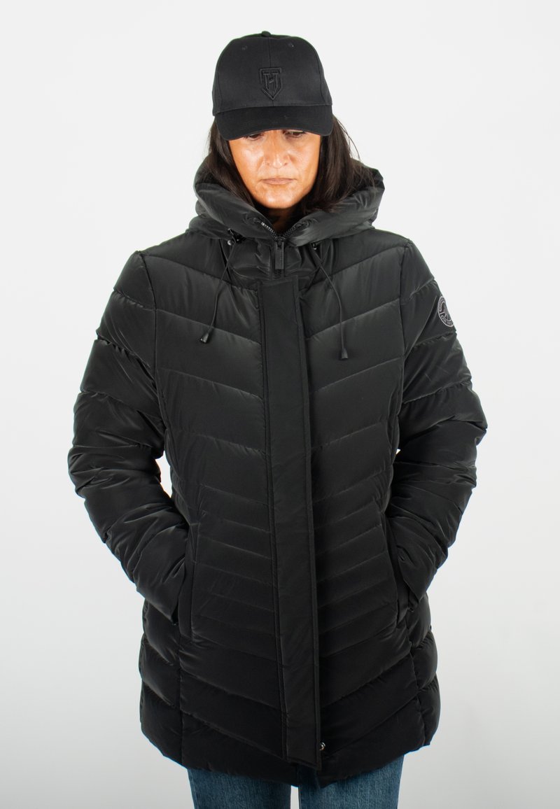 Doudoune noire avec capuche, présentant un design matelassé vertical, des poches latérales et des cordons réglables. Associée à une casquette noire.