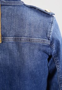 Tummansininen farkkutakki, jossa on teksturoidun pinnan lisäksi kultasävytetyt napit olkapäillä ja näkyvät saumat takana.