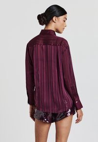 Frau mit einem Dutt, die eine transparente, gestreifte bordeauxrote Bluse und schimmernde, dunkelviolette Shorts trägt, steht vor einem schlichten, hellen Hintergrund.