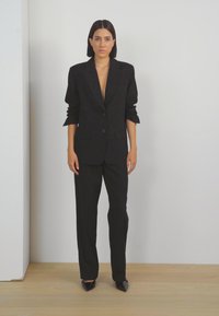 Černý přizpůsobený blazer s hlubokým výstřihem do V, ohrnutými rukávy a sladěnými kalhotami. Elegantní látka a boty s špičatou špičkou dotvářejí celkový vzhled.