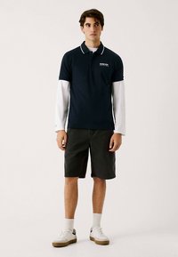 Marinblå pikétröja med vit krage, lager-på-lager över en vit långärmad skjorta. Matchas med mörka shorts och vita sneakers med bruna detaljer.