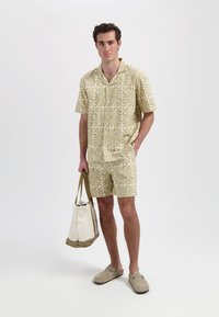 Beige shirt met korte mouwen en bijpassende shorts met een herhalend palmpatroon. Gecombineerd met lichtgrijze instap schoenen en een beige schoudertas.