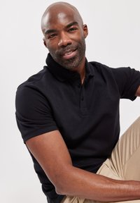 Zwarte polo met een gestructureerde stof, een placket met twee knopen en een klein logo-detail. Model poseert met lichtbruine broek.