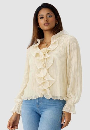 Donna che indossa una camicetta color crema a maniche lunghe con volant sul davanti e sui polsini, abbinata a jeans azzurri su sfondo semplice.