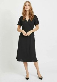 Černé zavinovací šaty s krátkými volánovými rukávy, délkou ke kolenům a jemnou texturou. Styling doplněn černými lodičkami se zlatými akcenty a špičatou špičkou.