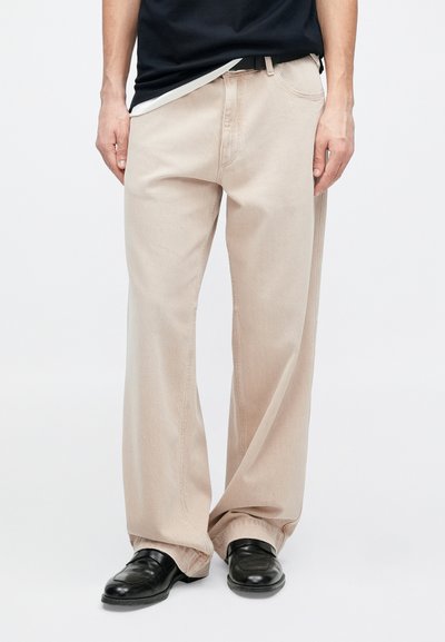 Μπεζ παντελόνι με φαρδύ πόδι και λεία υφή, σχέδιο με πέντε τσέπες και μαύρη ζώνη, σε συνδυασμό με μαύρα επίσημα παπούτσια.