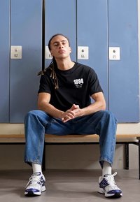 Tricou negru cu textul "1894", blugi albaștri, adidași albi și mov. Modelul este așezat pe o bancă în fața unor dulapuri albastre.