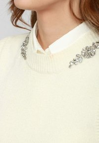 Maglione color crema con un tessuto a maglia testurizzato, caratterizzato da decorazioni in argento lungo il colletto sopra una camicia bianca.