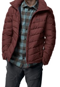 Homme portant une veste en duvet bordeaux dézippée sur une chemise à carreaux bleue et marron, avec un pantalon cargo gris foncé, les mains dans les poches.