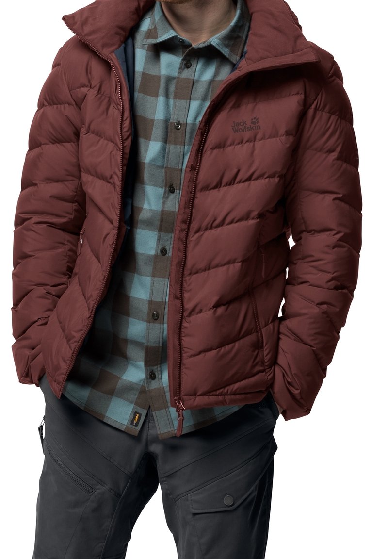 Homme portant une veste en duvet bordeaux dézippée sur une chemise à carreaux bleue et marron, avec un pantalon cargo gris foncé, les mains dans les poches.