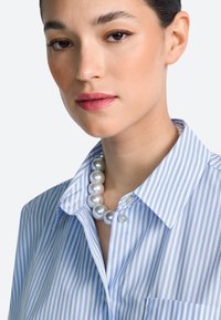 Modrá a biela pruhovaná košeľa s golierom, zdobená perlovým náhrdelníkom a detailom s gombíkom. Hladká tkanina s voľným strihom.