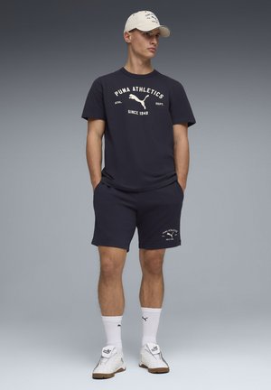 Marineblauw katoenen t-shirt en korte broek met wit Puma-logo en tekst; gecombineerd met witte sportsokken en sneakers, en een beige pet.