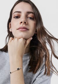 Bracciale in argento con accenti in forma di stella nera e fiore, caratterizzato da una delicata catena. Indossato su un polso, con una camicia grigia visibile sullo sfondo.
