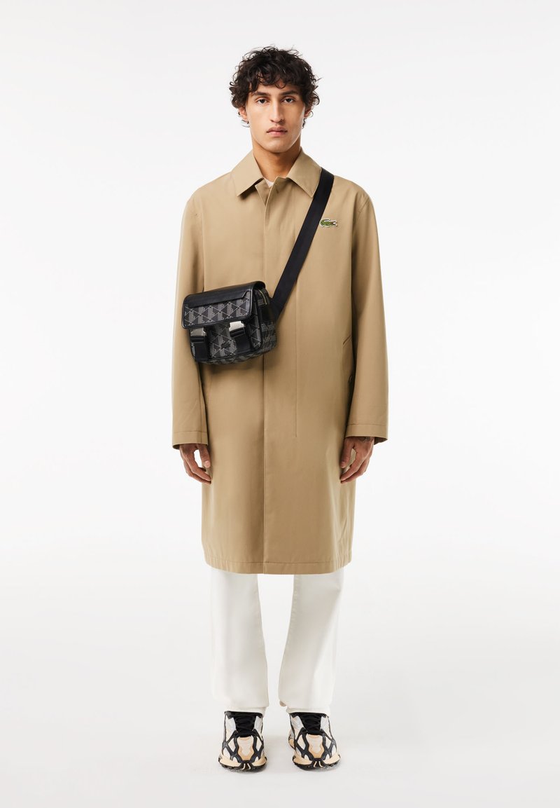 Beige Mantel mit Kragen, schwarze Tasche mit Muster und silbernen Beschlägen, weiße Hose und schwarze sowie cremefarbene Turnschuhe.