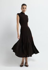 Hnědé žebrované pletené šaty s vysokým výstřihem a bezrukávovým designem. Rozšířená, skládána sukně sahá po střed lýtek a je doplněna hnědými botami.