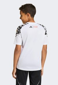 Hvid T-shirt med korte ærmer har et sort og gråt grafisk mønster på skuldrene, Adidas-logoen og en rød Marvel-mærkat på bagsiden af halsudskæringen.