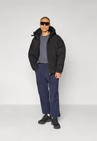 Άτομο με μαύρο μπουφάν, γκρι μπλούζα, μπλε cargo παντελόνι, μαύρα παπούτσια πεζοπορίας και μεγάλα μαύρα γυαλιά ηλίου στέκεται μπροστά σε έναν απλό τοίχο.