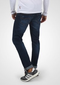 Mørkeblå denimjeans med en litt slitt design, med to baklommer og oppbrettede ermer, parret med grå idrettssko.