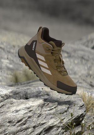Καφέ και μαύρο πεζοπορικό παπούτσι Adidas Terrex με λευκές ρίγες, εμφανίζεται στον αέρα πάνω από πετρώδες έδαφος.
