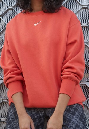 Osoba ubrana w jasnopomarańczową bluzę Nike z podwiniętymi rękawami, siedząca na tle perforowanej metalowej ściany.