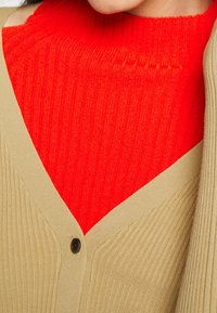 Cardigan beige côtelé avec un col en V et un bouton noir unique, superposé à un col roulé côtelé rouge vif.