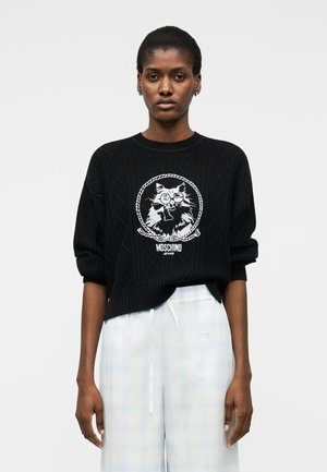 Ženska, oblečena v črn pleten pulover z grafiko mačke in napisom "Moschino Jeans", v kombinaciji s svetlo modrimi in belimi karo hlačami.