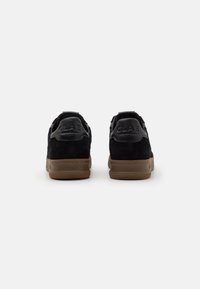 Black suede sneakers with rubber soles. Features embossed branding on the heel and a textured design. Rear view highlighted.