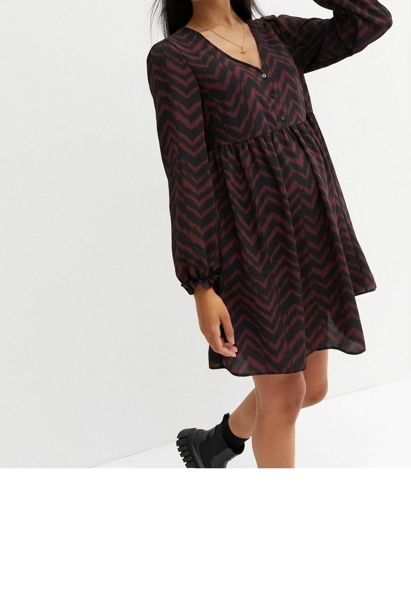 Femme portant une robe à manches longues noire et rouge foncé à motif zigzag avec des boutons et des bottines, posant devant un fond blanc uni.