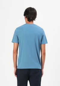 Bărbat cu părul închis la culoare, purtând un tricou simplu albastru cu mânecă scurtă și pantaloni negri, stând cu spatele la un fundal alb.