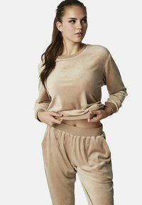 Beige velours loungewear-set bestaande uit een langemouwen top en broeken met een elastische tailleband en zijzakken. Zachte textuur en gelegenheidsfit.