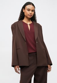 Woman with long curly hair wearing a loose brown blazer, matching pants, and a burgundy top with a keyhole neckline and gold accent.