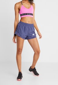 Vaaleanpunainen urheiluliivi, jossa on musta Nike Air -nauha, yhdistetty tummiin purppurashortseihin, joissa on valkoinen logo. Mallilla on päällä mustat urheilukengät, joissa on vaaleanpunaiset pohjat.