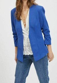 Blå kavaj med en strukturerad design, långa ärmar och ett mjukt material. Lager ovanpå en vit topp med spetsdetaljer, matchad med jeans.