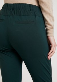 Pantalons élastiques couleur bleu sarcelle avec une texture lisse, dotés d'une taille plate et de poches arrière. Design simple et ajusté.