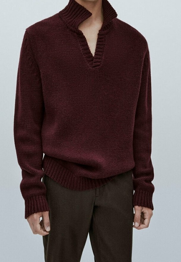 Personne portant un pull en maille bordeaux foncé avec un col en V profond et un col haut, associé à un pantalon brun foncé, debout devant un fond uni.