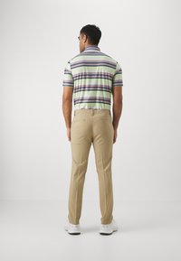 Camisa polo a rayas en colores verde, púrpura y blanco, combinada con pantalones beiges. El atuendo incluye zapatos deportivos blancos, mostrados desde atrás.