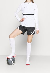 Weibliche Fußballspielerin in weißem Langarmshirt, schwarzen Shorts, weißen Knieho socks und weißen Fussballschuhen, mit einem Fuß auf einem schwarz-weißen Fußball.