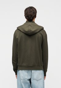 Φούτερ με κουκούλα σε ελιά πράσινο, με υφασμάτινο πλεκτό σχέδιο, που διαθέτει πίσω πατιλέτα, μεγάλη κουκούλα και ριμπ μανσέτες και τελείωμα. Φορεμένο πάνω από ανοιχτόχρωμο μπλε τζιν.