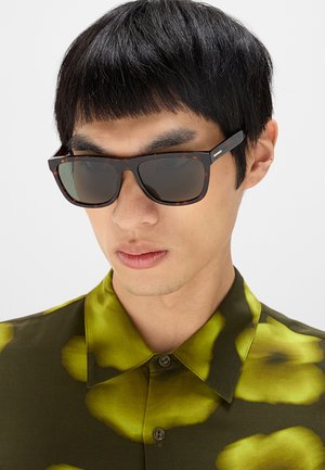 Giovane uomo con capelli neri corti che indossa occhiali da sole rettangolari scuri e una camicia con bottoni a fantasia verde-gialla.